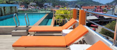 A rooftop pool area with orange lounge chairs, a swimming pool, and a view of a town with buildings and hills in the background.