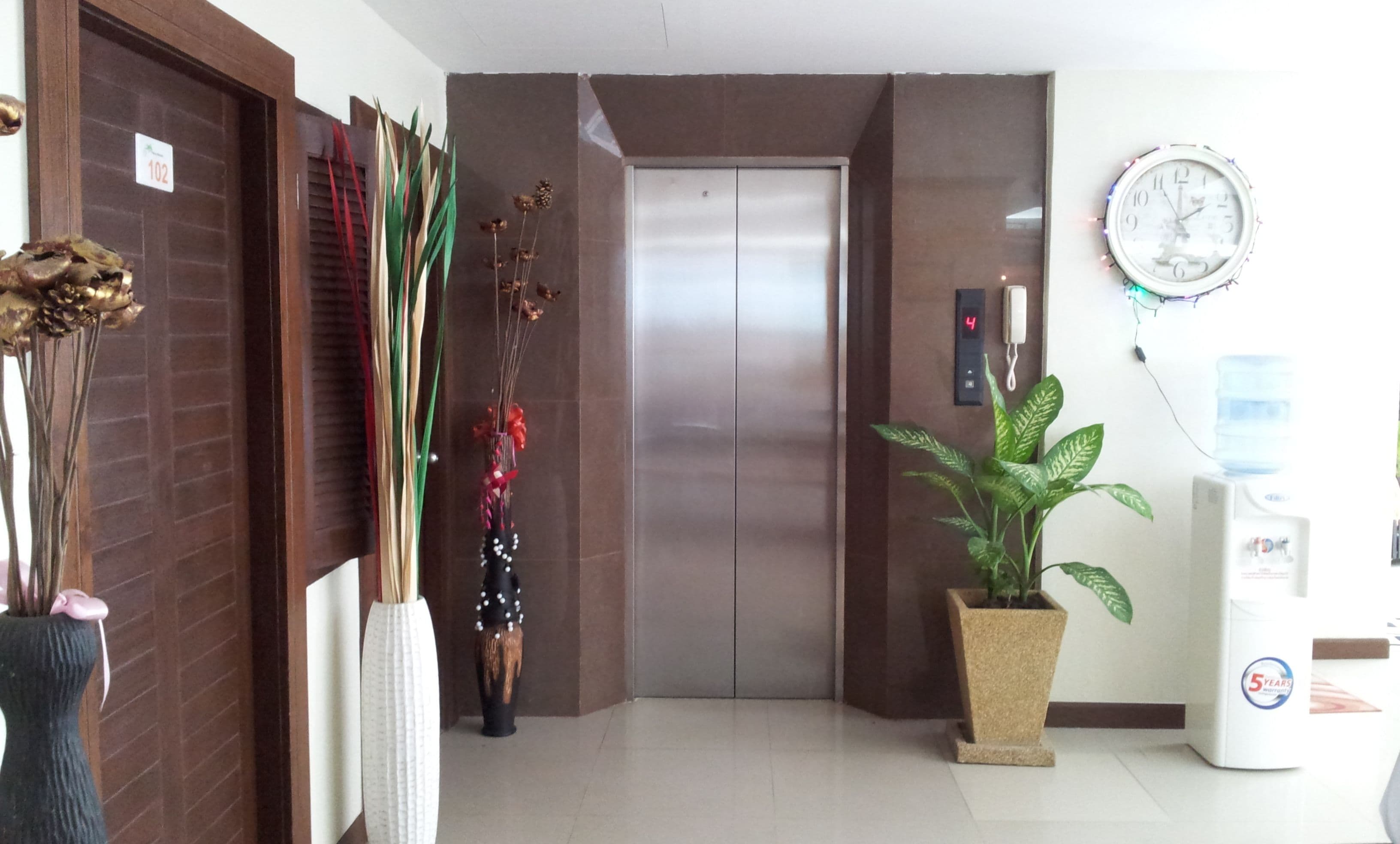 Un pasillo con una puerta marcada con el número 102, un dispensador de agua, un reloj y una planta en maceta.