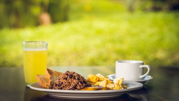 A breakfast setting with a plate of food, a glass of juice, and a cup on a table outdoors. - Brisas Arenal Hotel (La Fortuna)