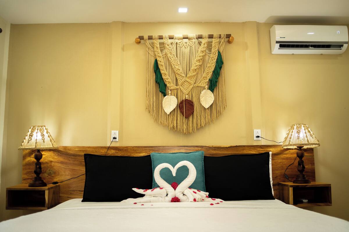 A hotel room with a bed, two bedside lamps, a wall-mounted air conditioner, and a decorative macramé piece above the bed.