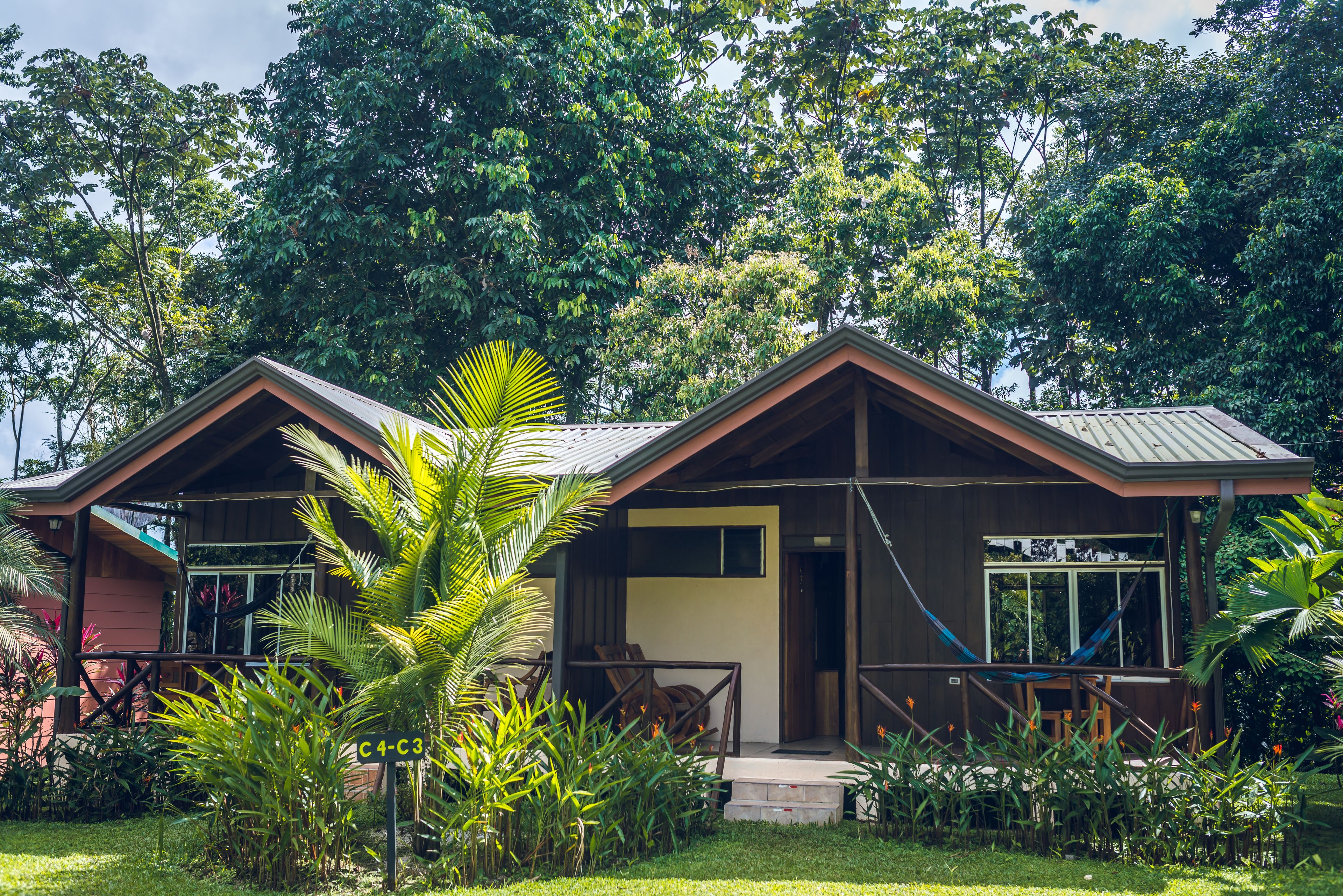 A two-story wooden house with a porch, surrounded by lush greenery.