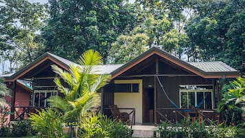 A two-story wooden house with a porch, surrounded by lush greenery.