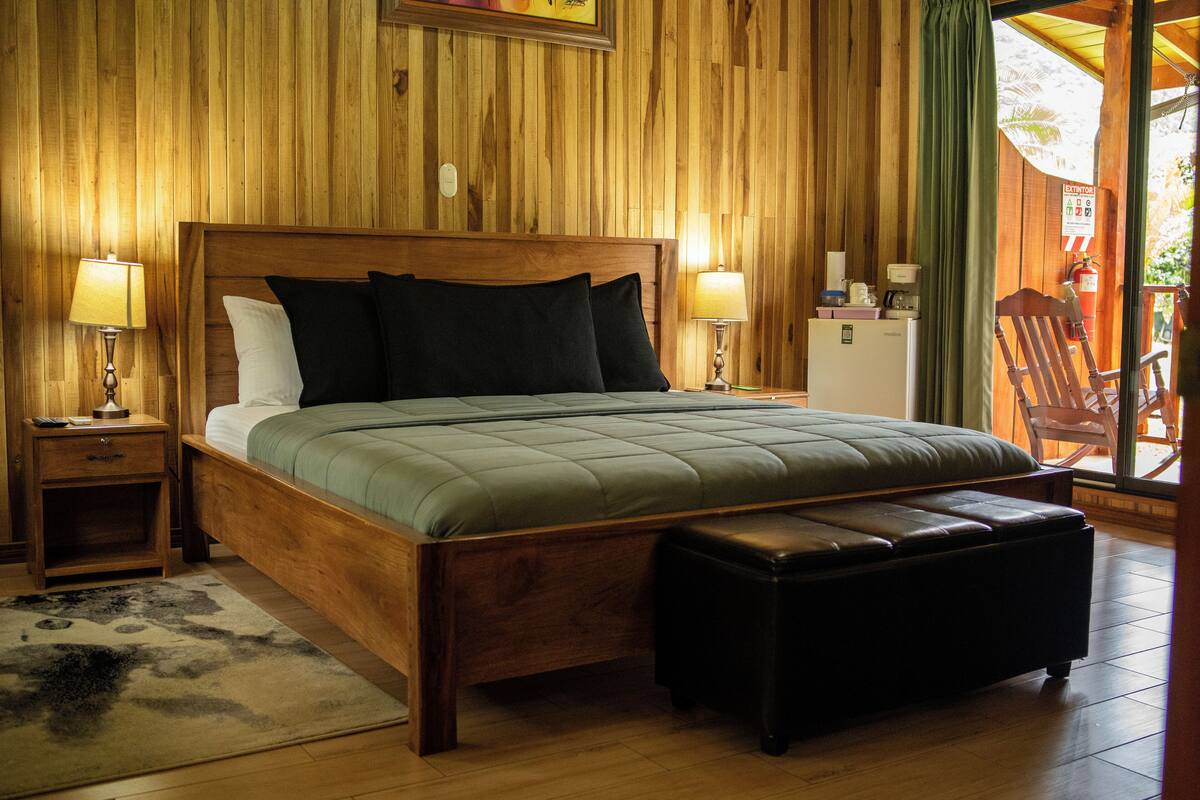 A wooden-paneled bedroom with a large bed, bedside lamps, a nightstand, a small refrigerator, and a wooden chair.