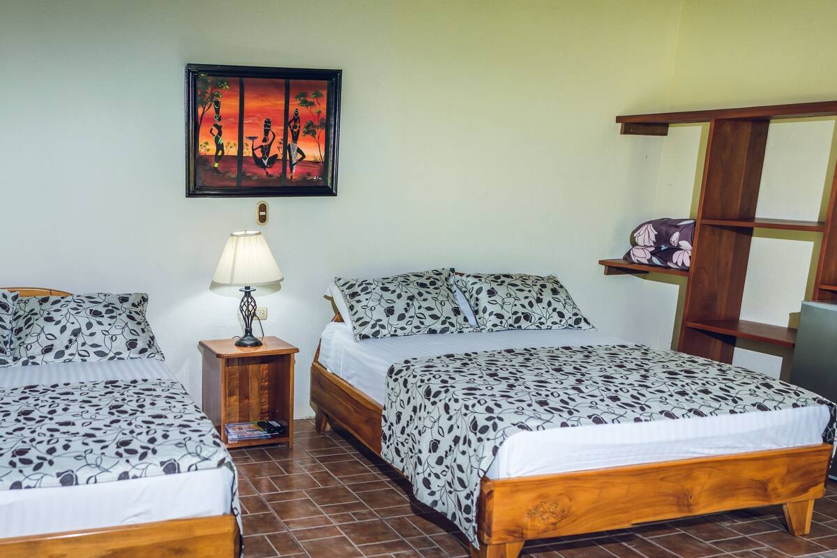 A room with two beds, a desk, a chair, a lamp, a refrigerator, and a painting on the wall.