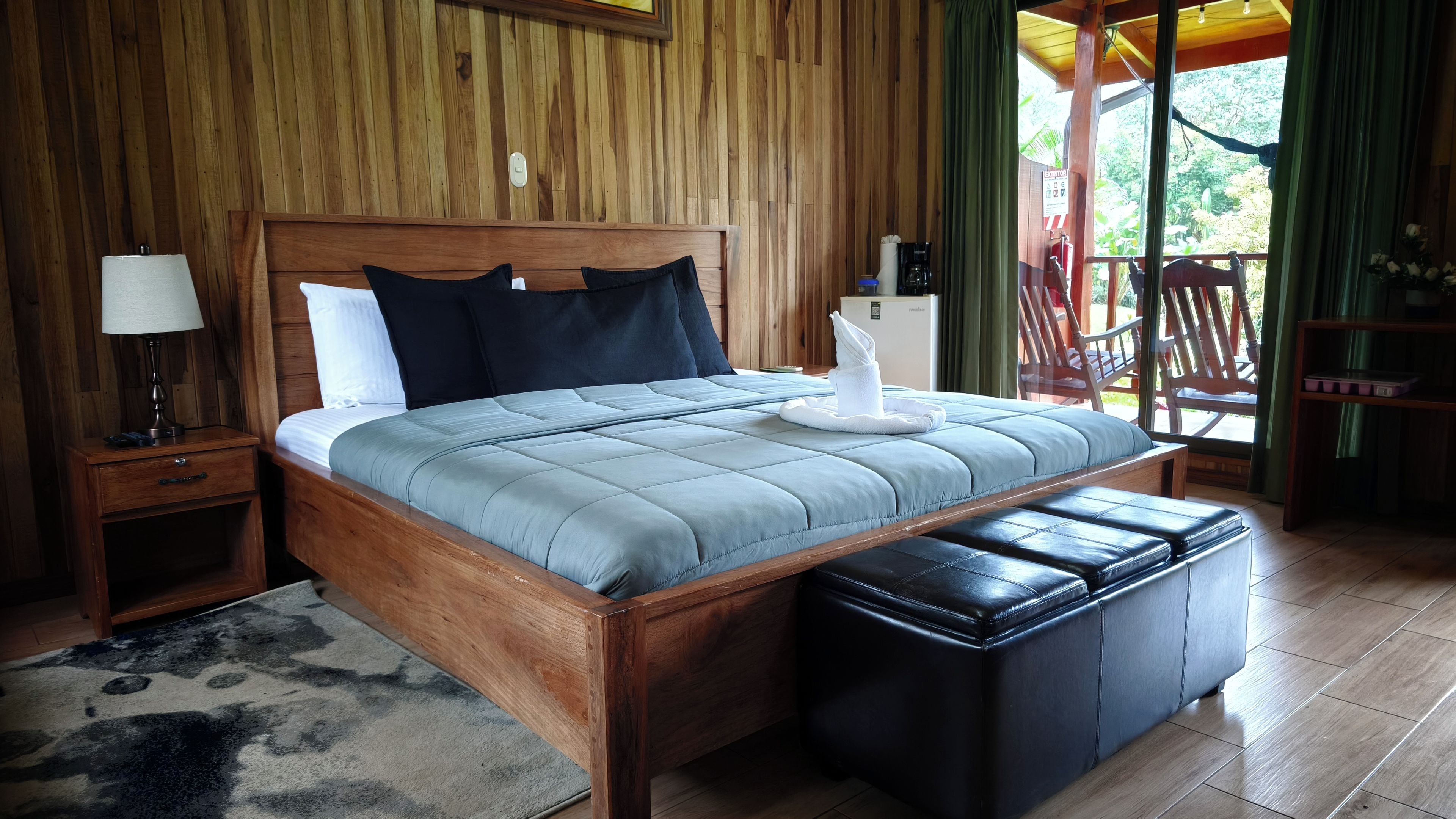 A wooden-framed bed with a grey quilt, a bedside lamp, and a small nightstand with a drawer.