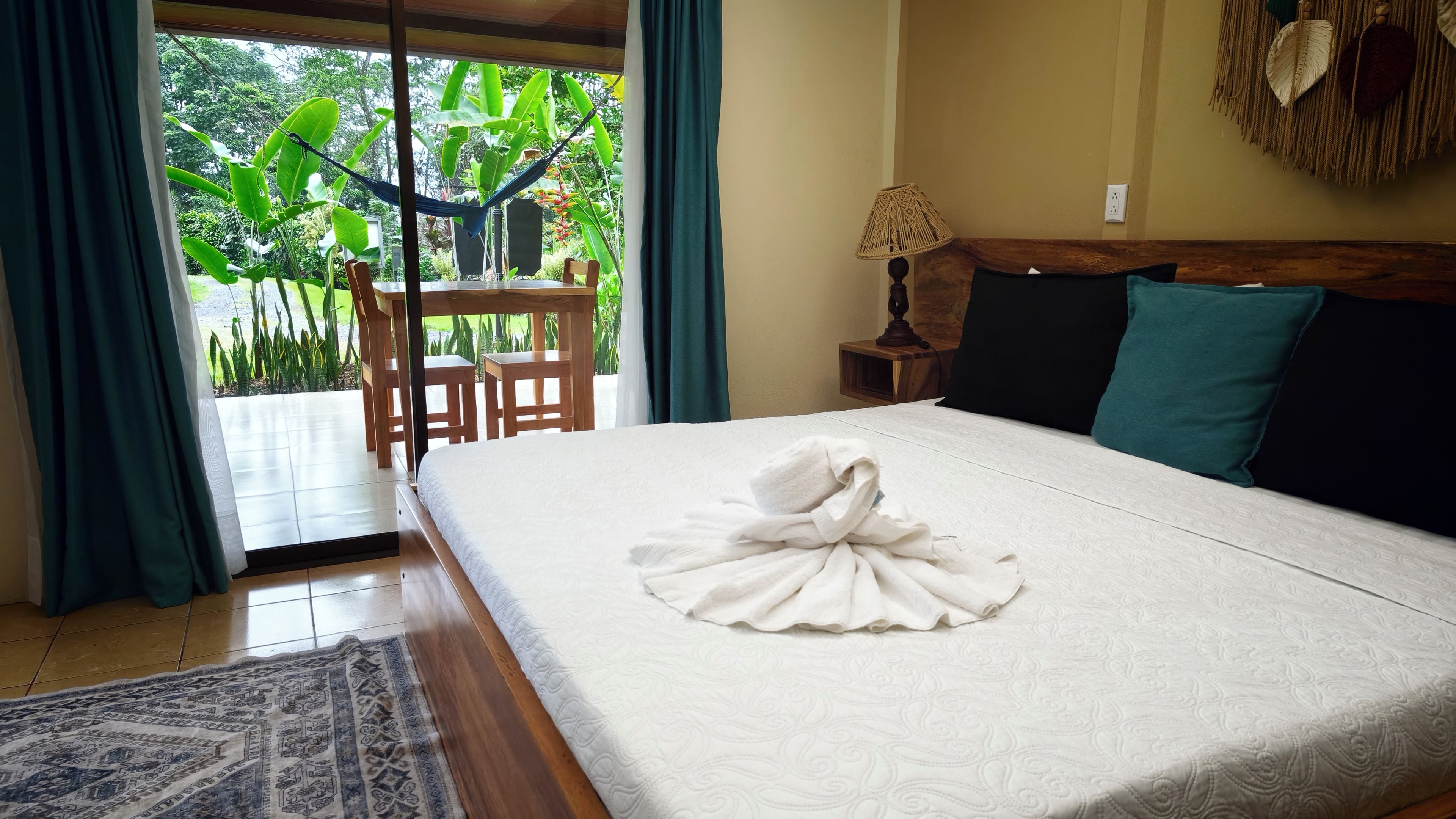 A hotel room with a bed, a wooden headboard, a nightstand, a lamp, and a view of greenery through a window.