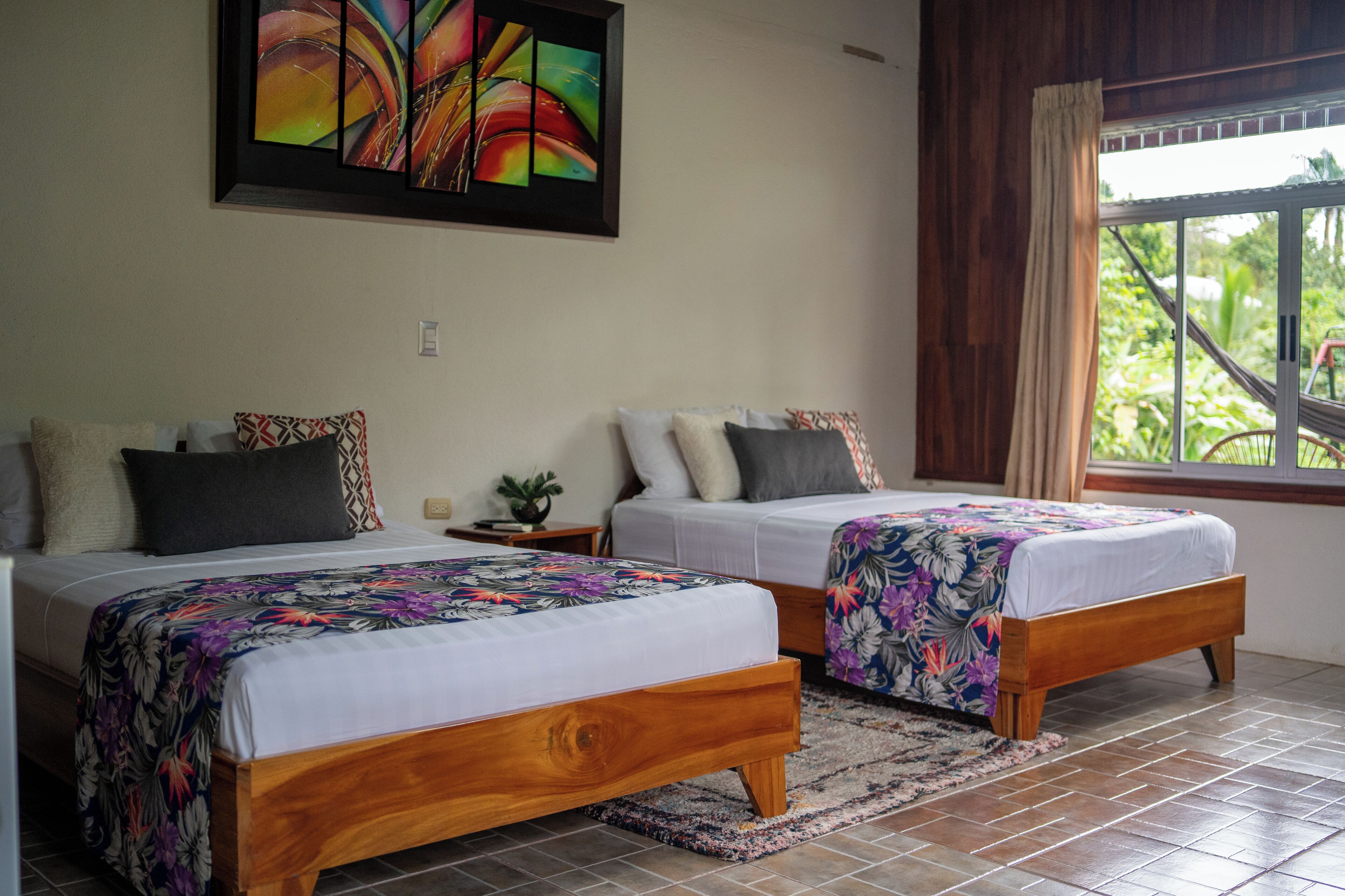 A room with two wooden beds, each with a floral bedspread, a large abstract painting on the wall, and a window with a view of greenery.
