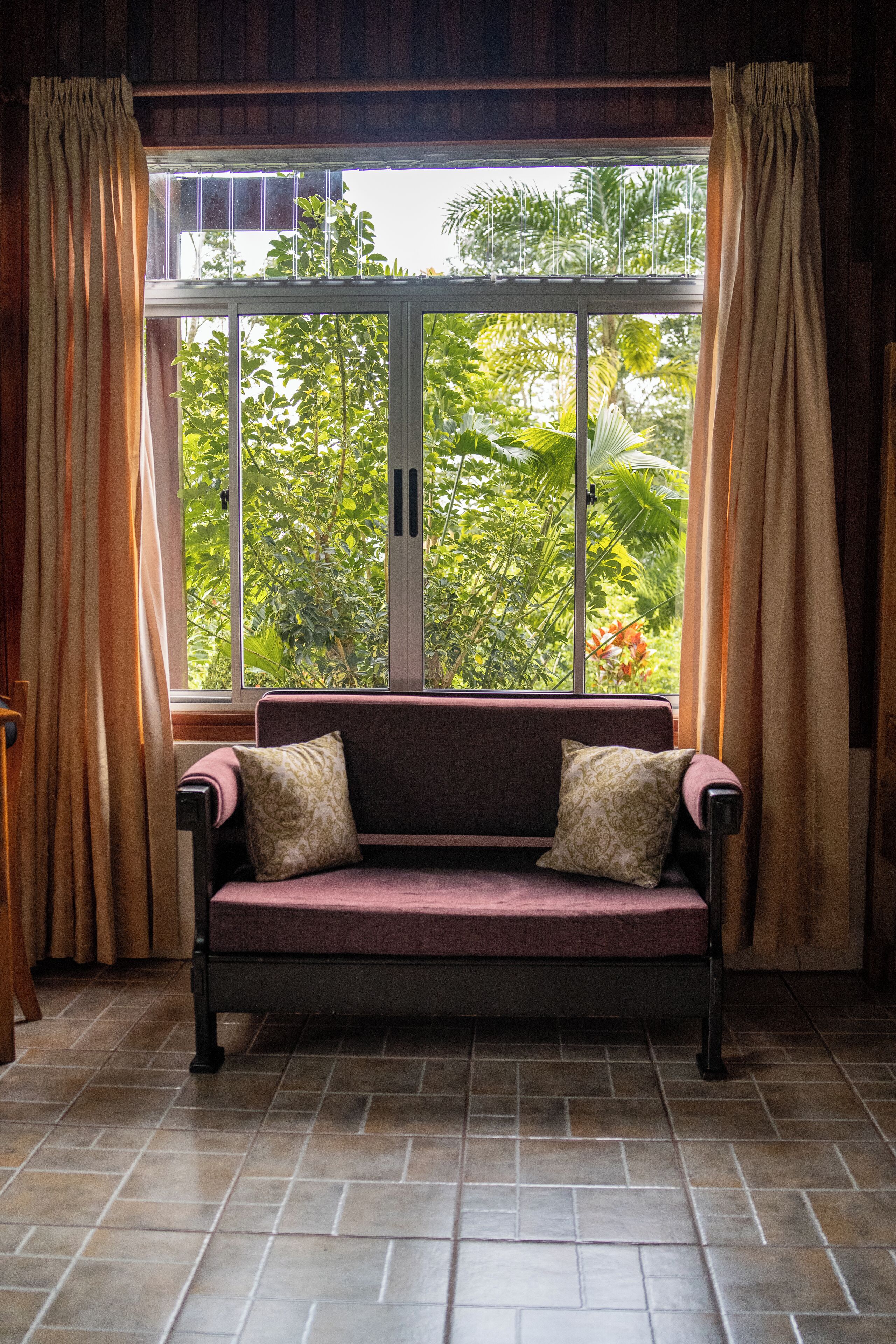 A room with a sofa facing a window with curtains, a tiled floor, and a view of greenery outside.