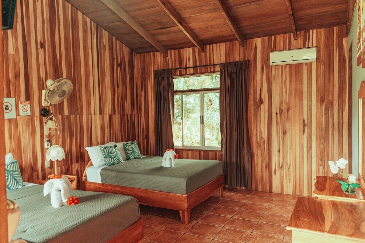 A wooden room with a bed, a small table, a fan, and a window.