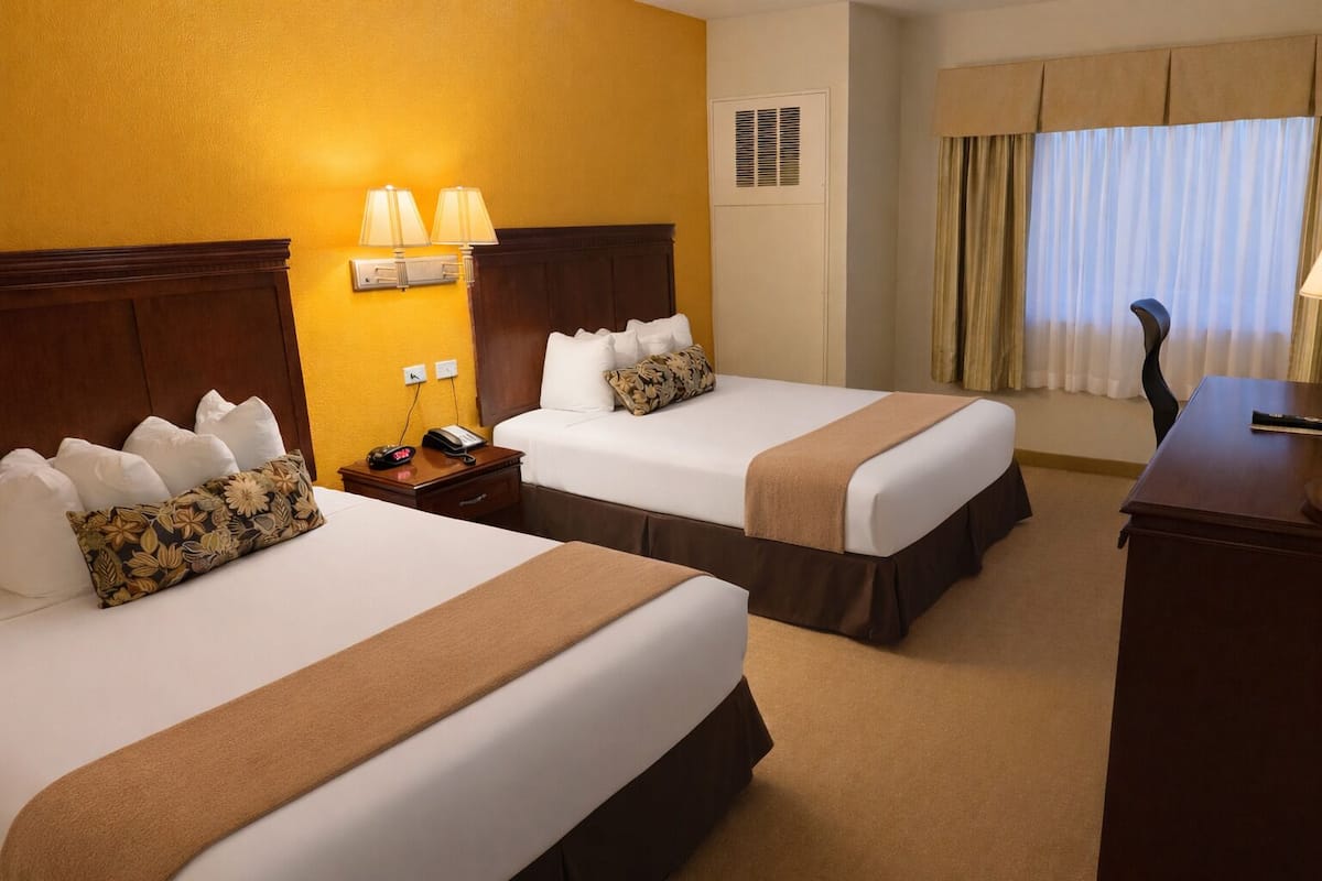 A hotel room with two beds, each with white linens and brown headboards. There are two bedside lamps, a nightstand with a phone and a lamp, and a desk with a lamp. The room has a window with curtains and a vent on the wall.