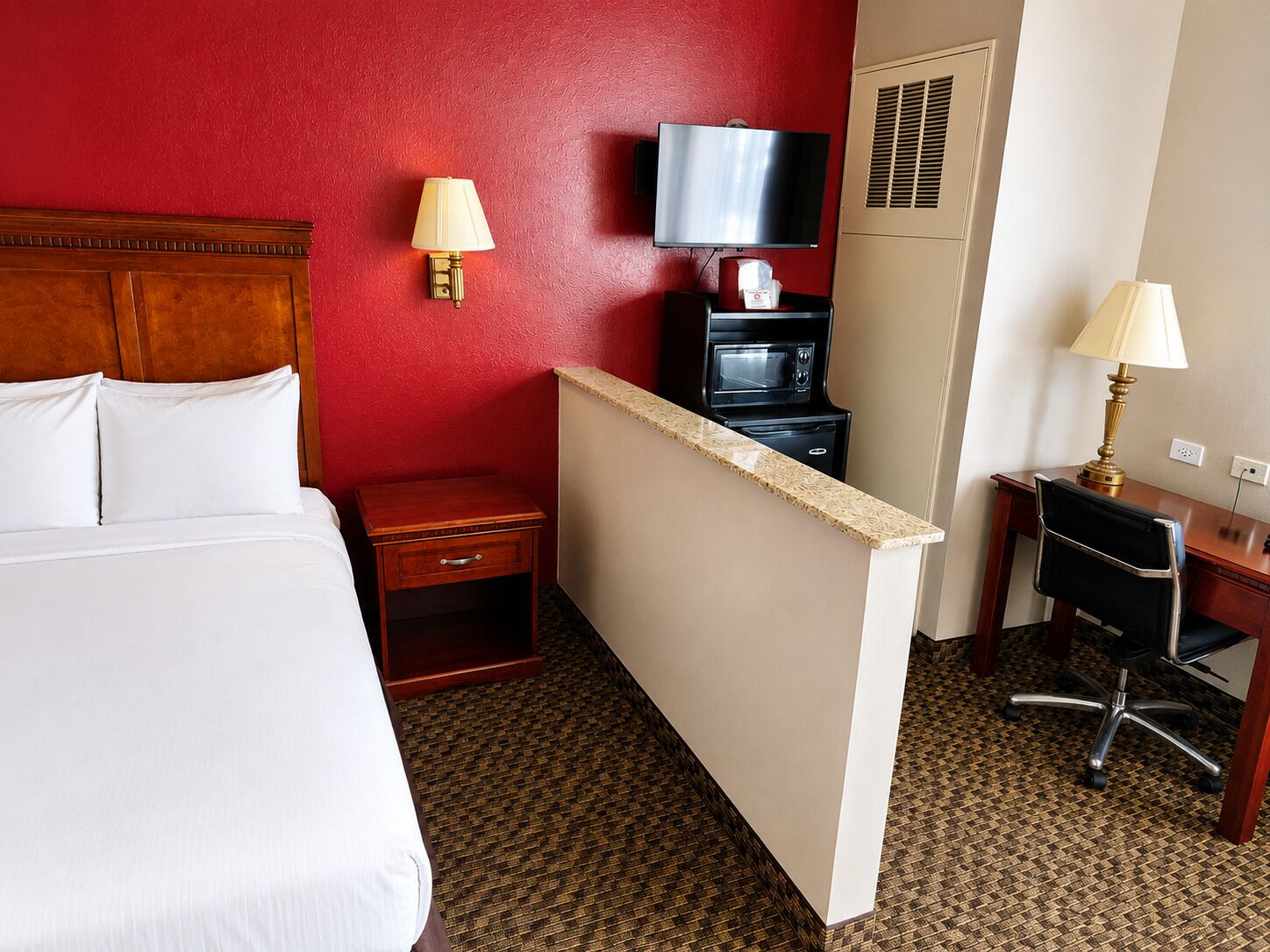 A hotel room with a bed, a desk, a television, a microwave, and a red wall.