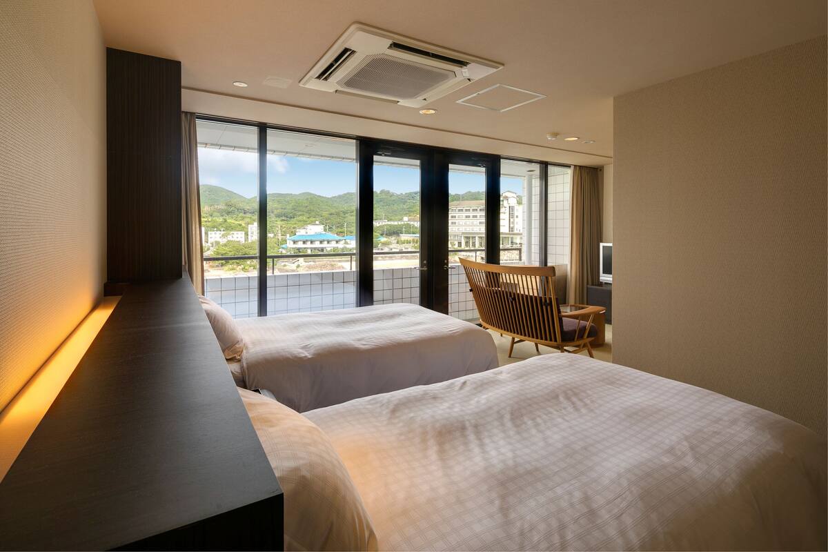 A hotel room with two beds, a large window with a city view, a chair, and a TV.