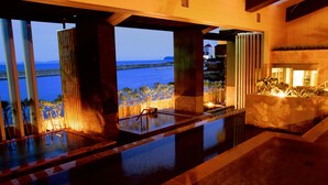 A luxurious indoor pool area with a view of the ocean, featuring a stone-tiled floor, wooden lounge chairs, and a fire feature. - Shimahana (Sumoto)