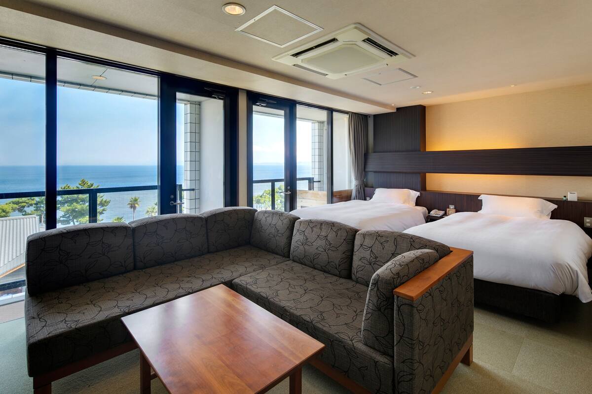 A hotel room with a sofa, two beds, a wooden table, and large windows overlooking the sea.