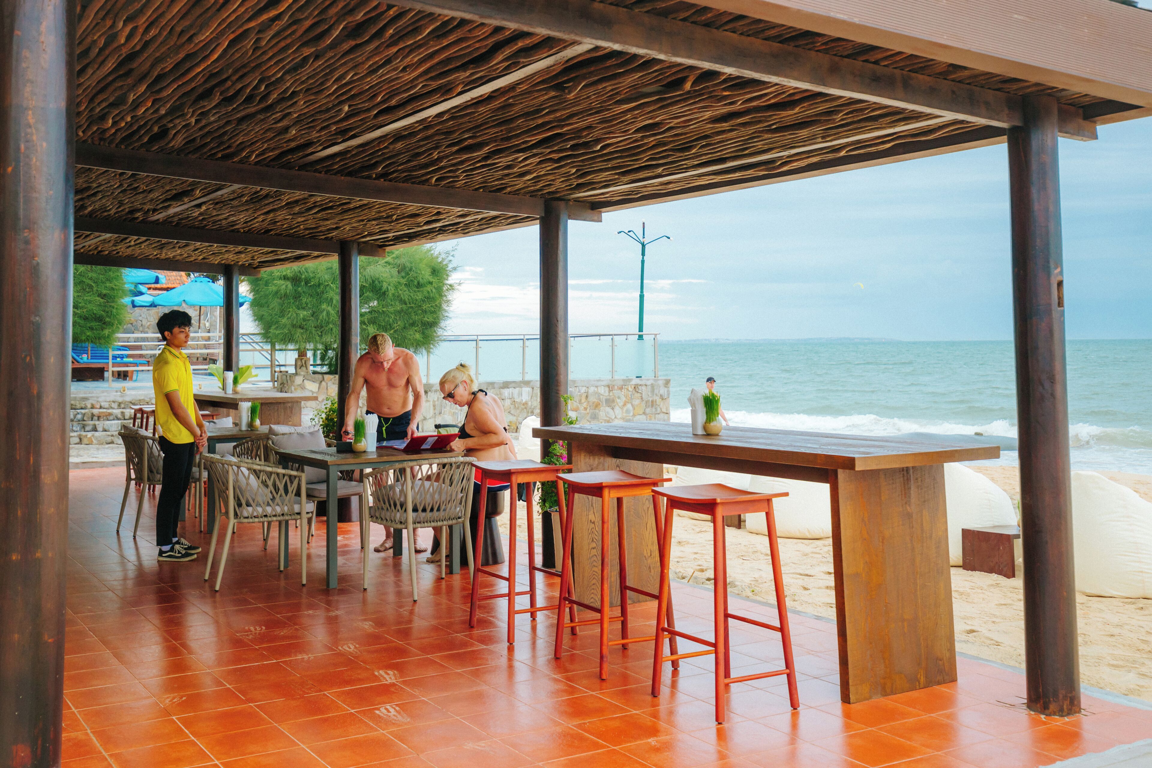 An outdoor seating area with a wooden pergola, tables, and chairs, overlooking the beach and sea.