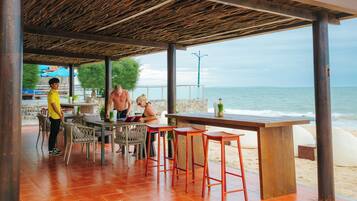 An outdoor seating area with a wooden pergola, tables, and chairs, overlooking the beach and sea.
