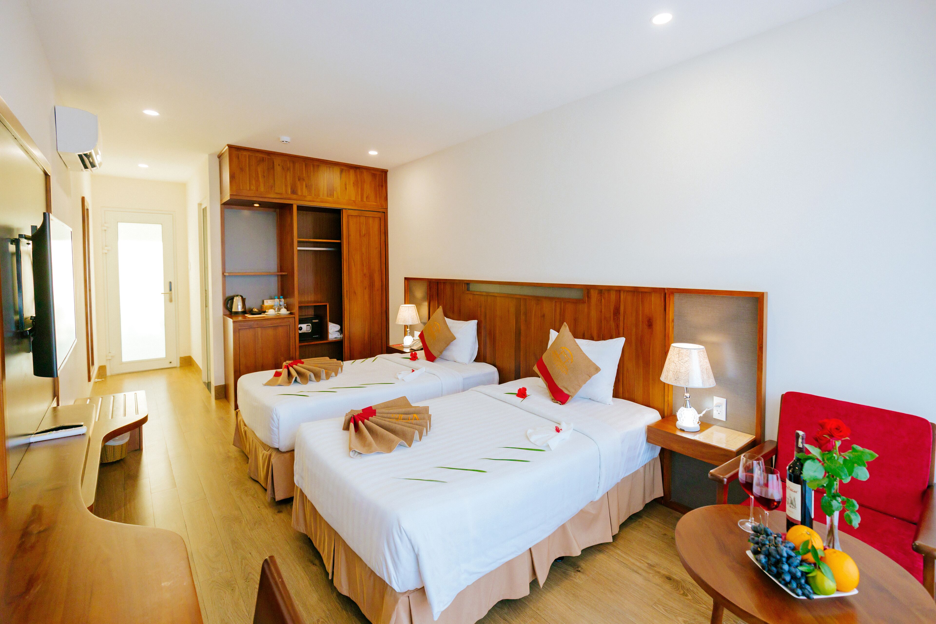 Habitación de hotel con dos camas, un escritorio, una silla, una mesita con fruta y vino, y un sillón rojo.