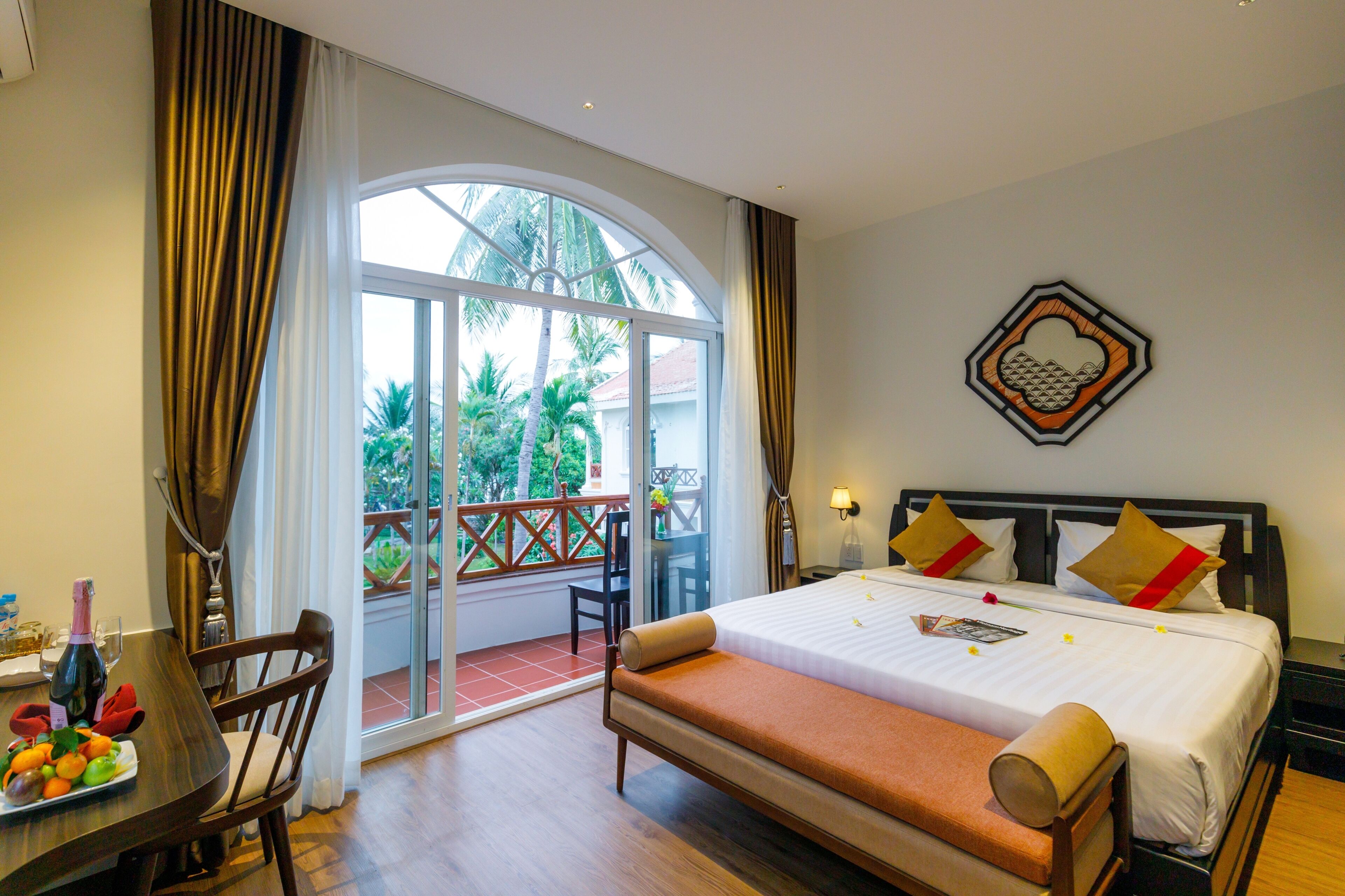Habitación de hotel con cama, mesa de comedor con sillas, balcón con vista a palmeras y un adorno decorativo en la pared.