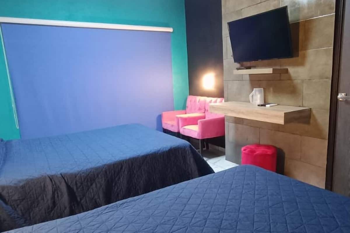 A room with two beds, a pink chair, a TV mounted on the wall, and a small table with a cup.