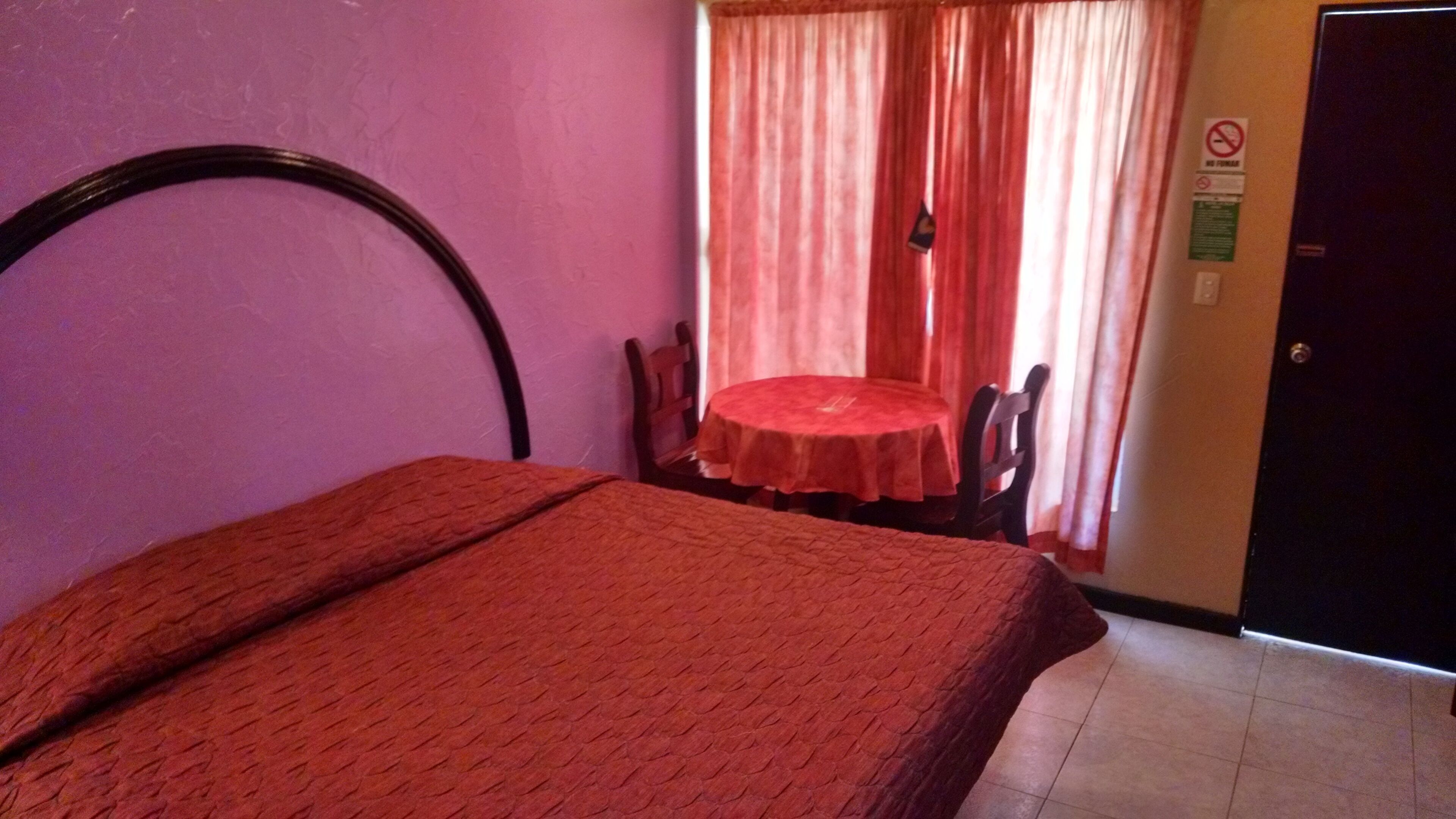 A room with a bed, a small table, and a chair. There is a door and a window with curtains.