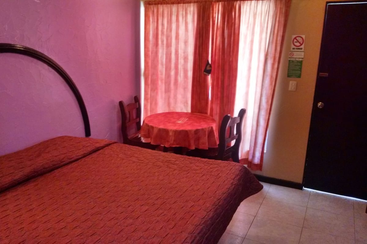 A room with a bed, a small table, and a chair. There is a door and a window with curtains.