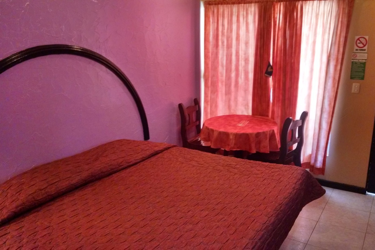 A room with a bed, a small table, and a chair. There is a door and a window with curtains.