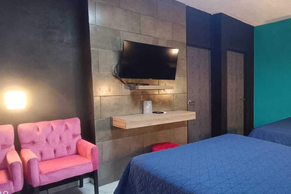 A hotel room with two beds, a TV mounted on a wall, a pink chair, and a teal accent wall.