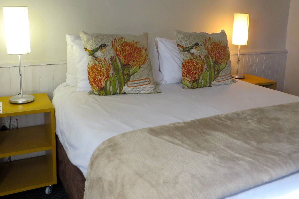Een net opgemaakt bed met witte lakens en twee decoratieve kussens met vogel- en bloemmotieven. Op gele nachtkastjes staan twee bedlampjes.