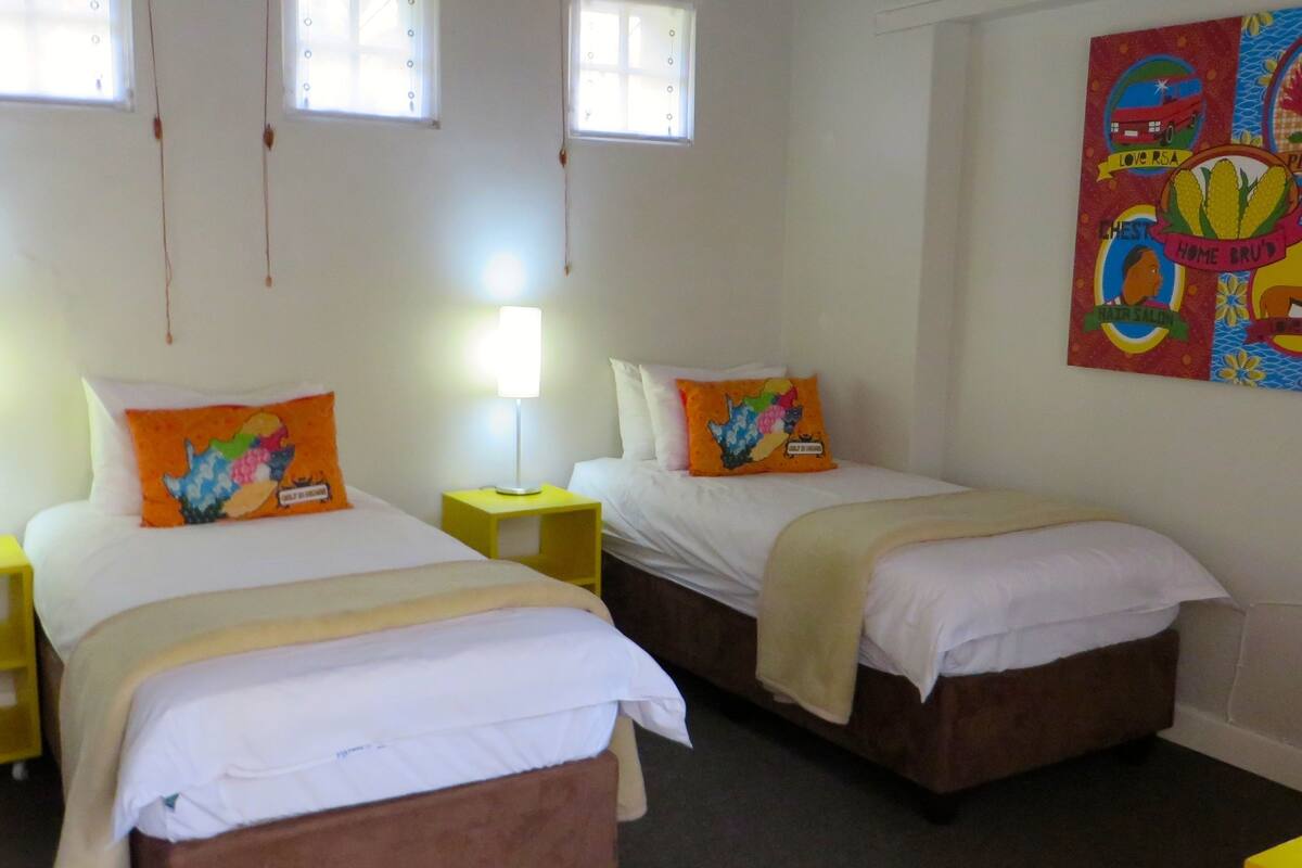 Een hotelkamer met twee bedden, een nachtkastje, een lamp en een kleurrijke wanddecoratie.