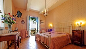 Quarto com cama, mesa de cabeceira e um pequeno armário. Há uma janela com vista para vegetação lá fora.