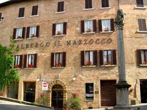 A stone building with the sign "Albergo Il Marzocco" indicating it is a hotel. There are multiple windows with shutters and flower boxes.