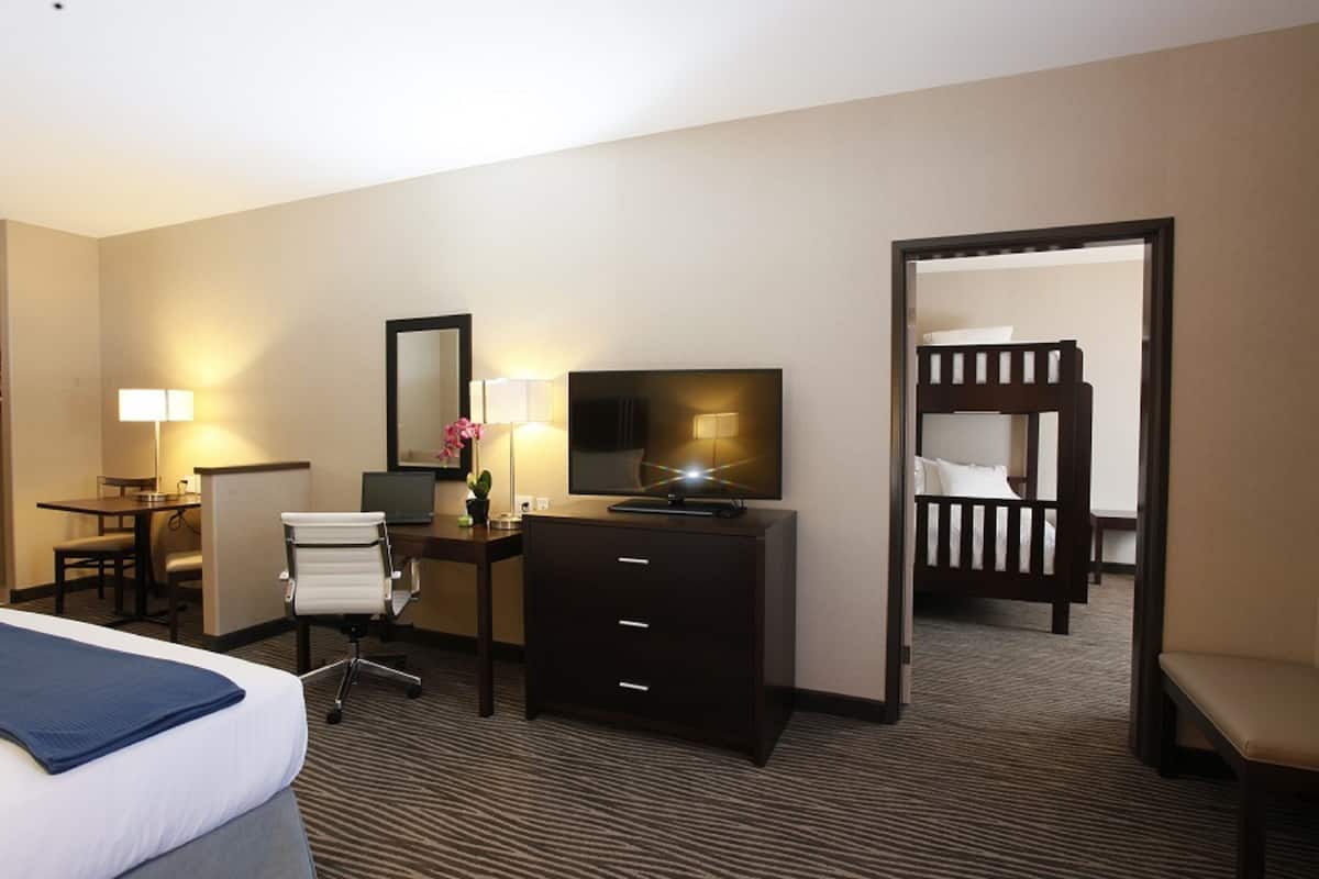 A hotel room with a bed, desk, chair, dresser, TV, and a bunk bed in the adjacent room.
