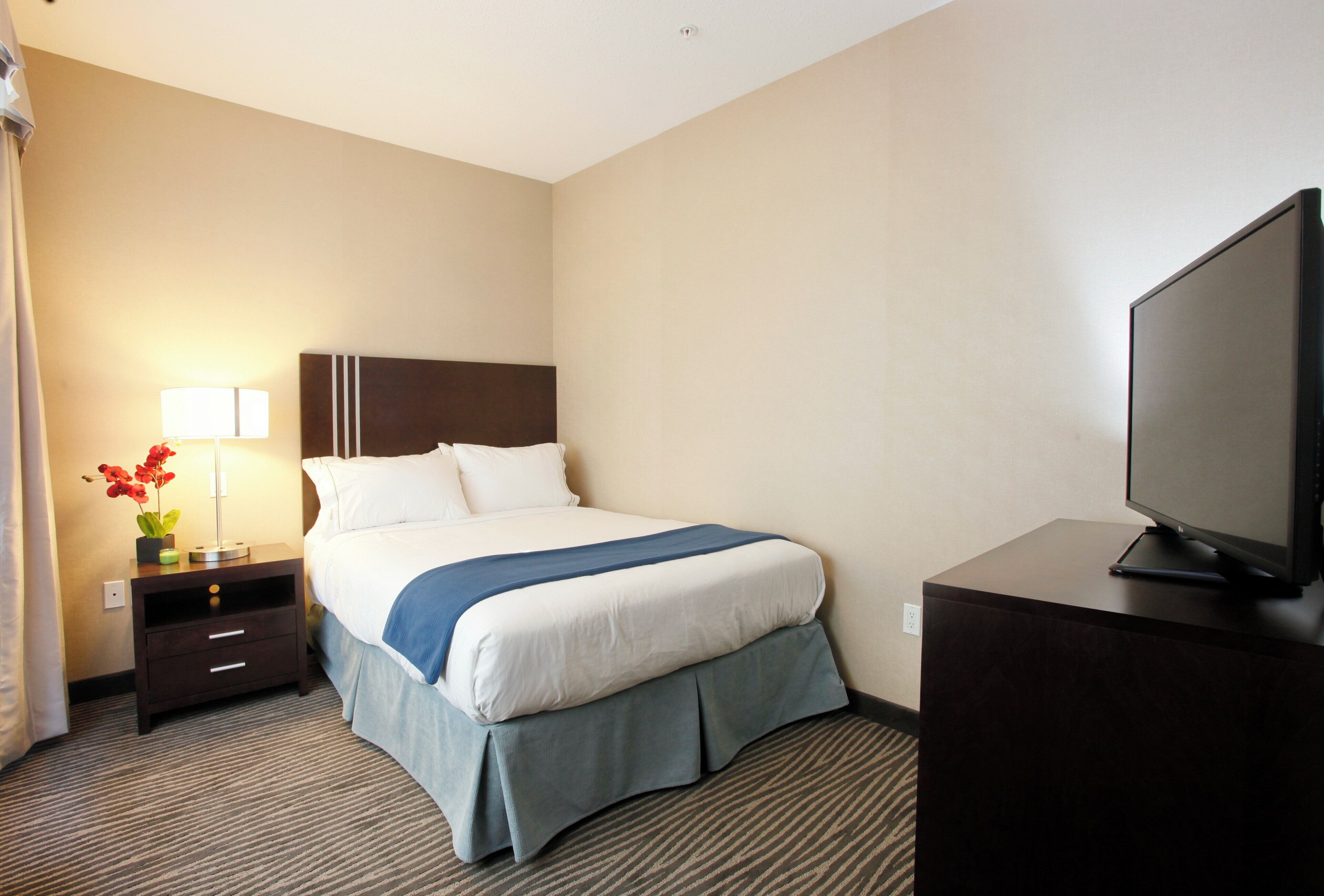 A hotel room with a bed, a desk, a television, and a nightstand with a lamp and flowers.