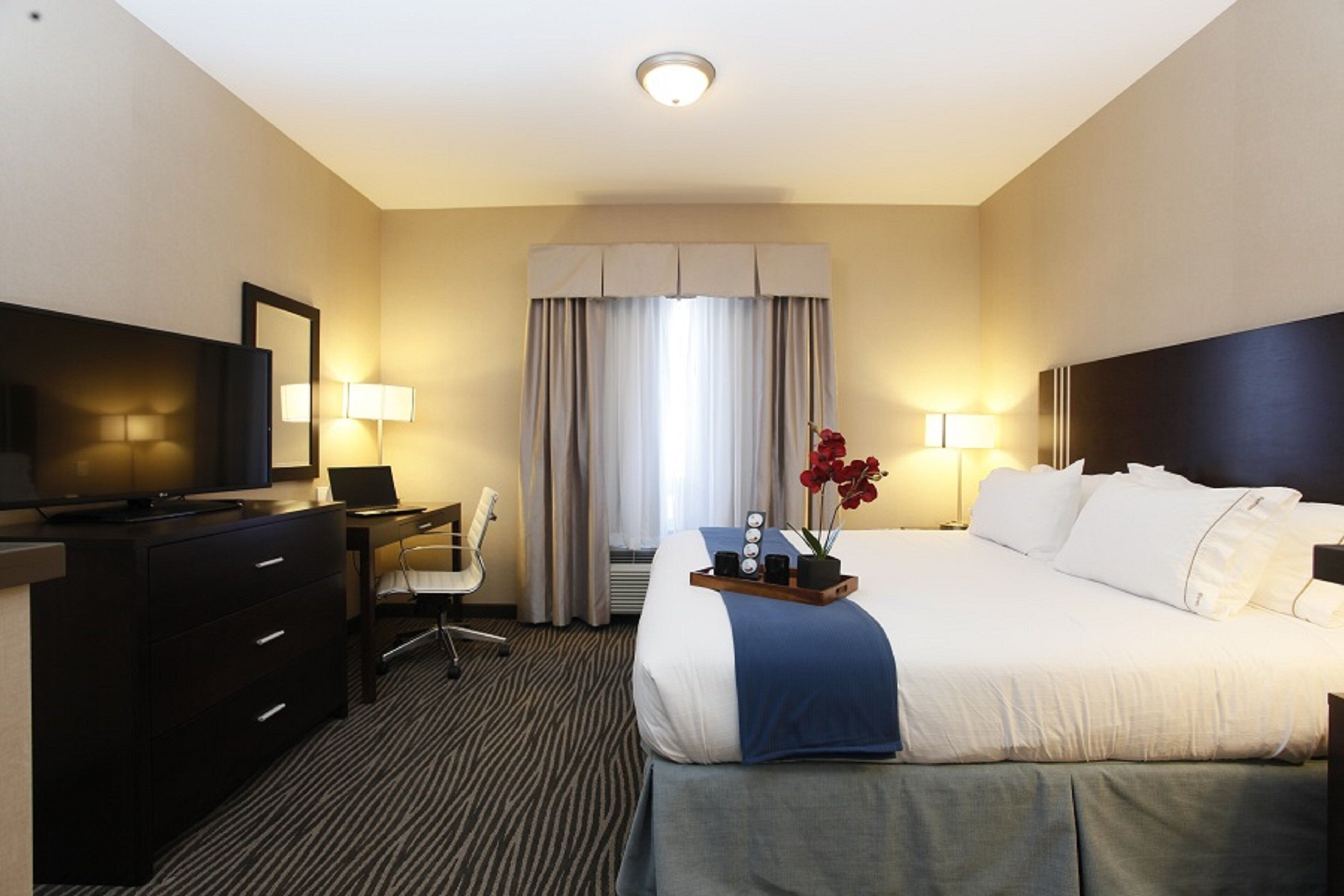 Quarto de hotel com cama, escrivaninha, cadeira, televisão e janela com cortinas.