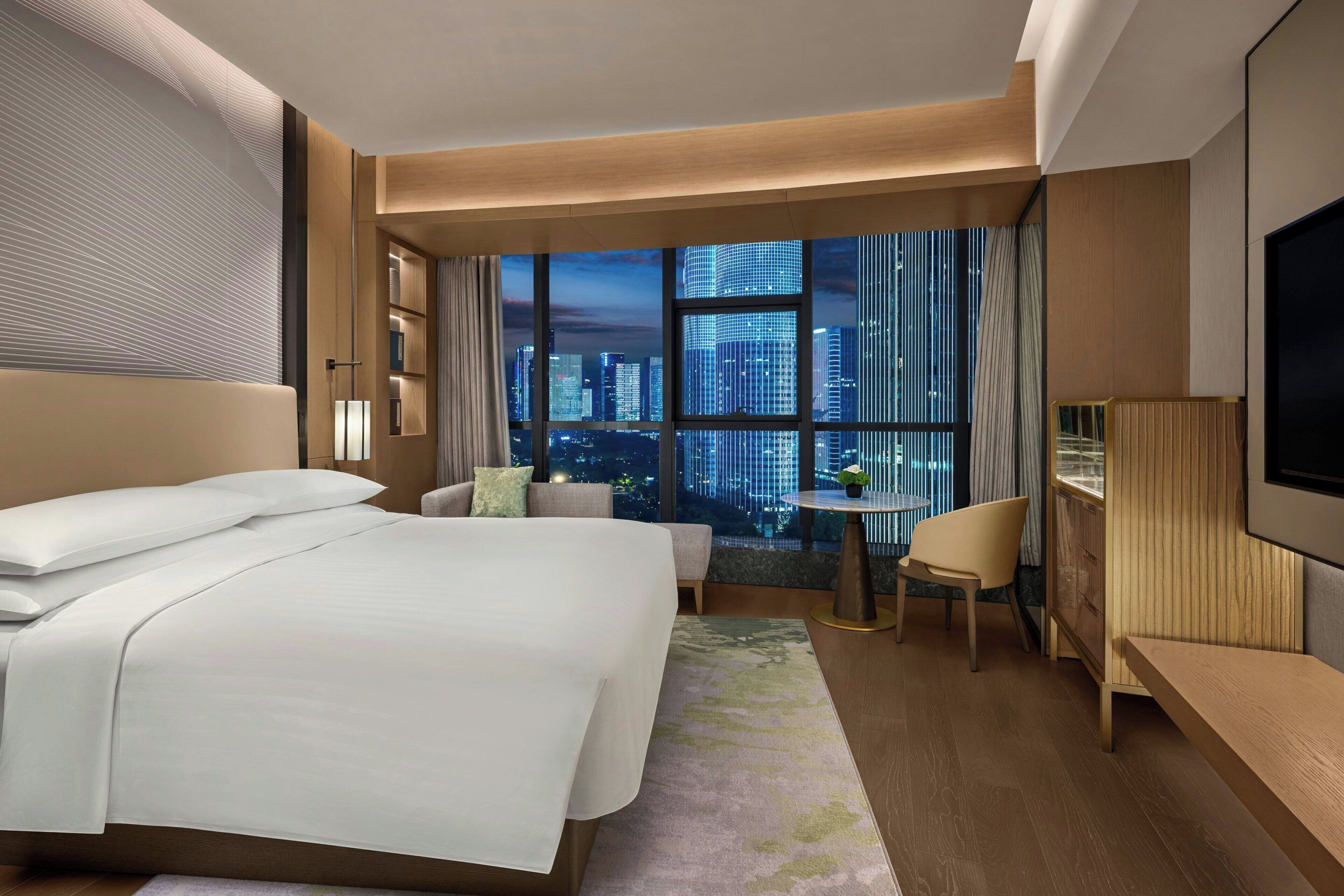 Ein Hotelzimmer mit einem großen Bett, einem Schreibtisch, einem Sessel und Blick auf die nächtliche Skyline.