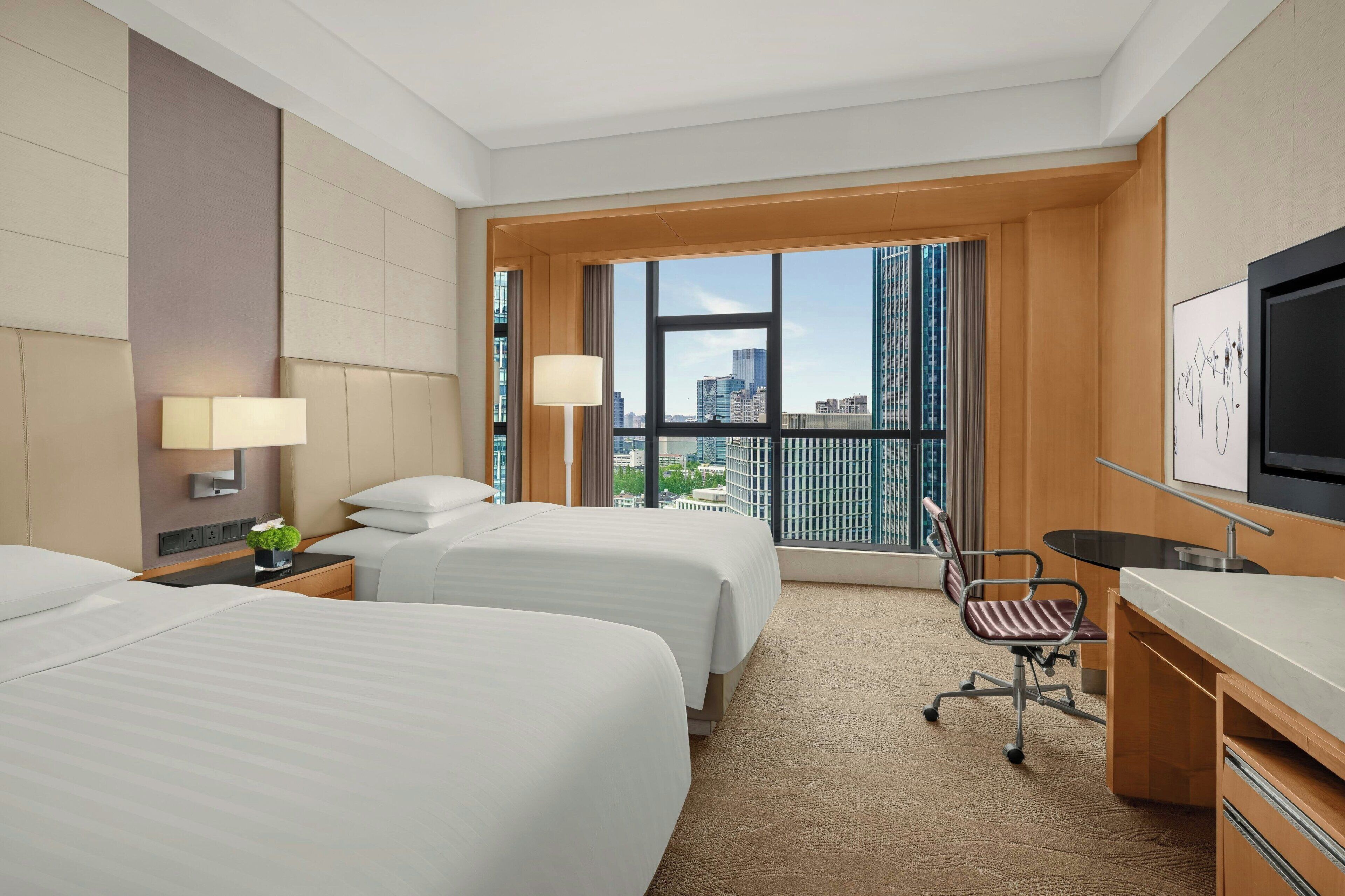 A hotel room with two beds, a desk, a chair, and a large window offering a city view.