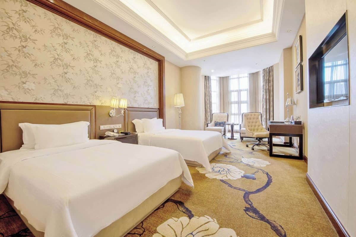 A hotel room with two beds, a desk, a chair, and a large window.