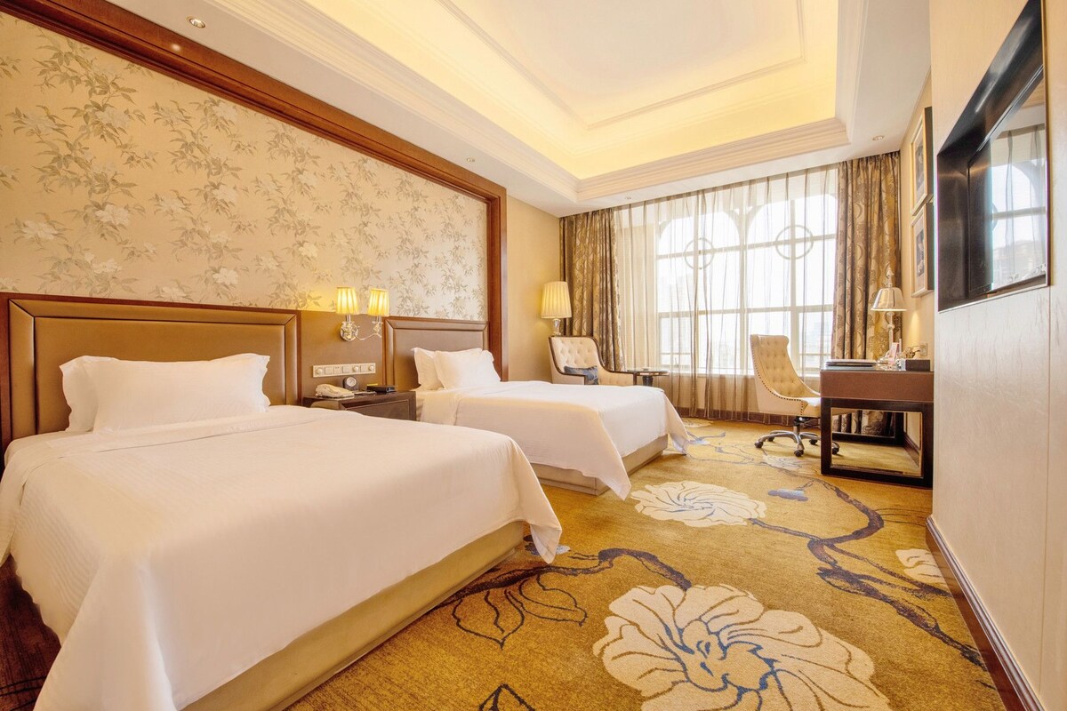 A hotel room with two beds, a desk, a chair, and a large window with curtains.