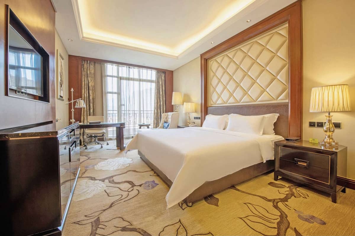 A hotel room with a large bed, a desk, a chair, a lamp, and a large window.