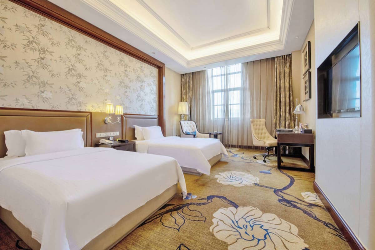 A hotel room with two beds, a desk, a chair, a TV, and a large window with curtains.