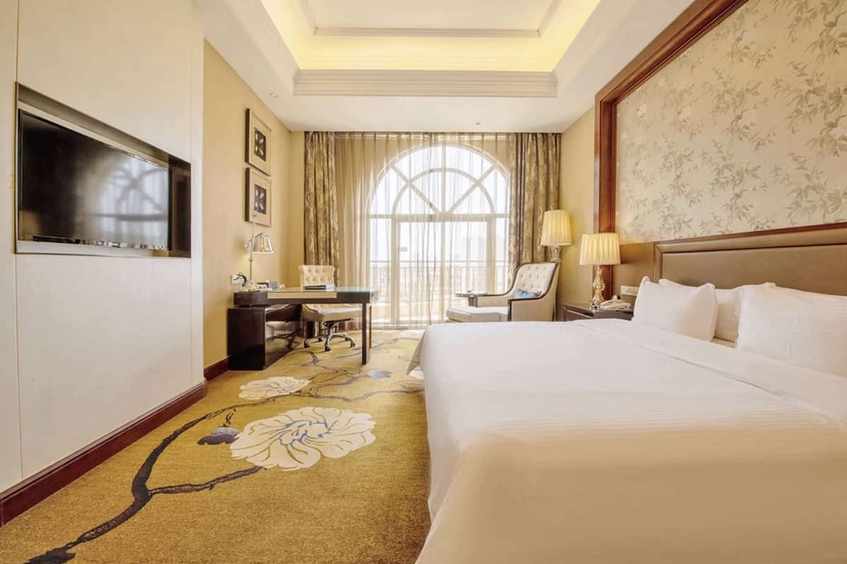 A hotel room with a large bed, a desk with a chair, a TV mounted on the wall, and a large window with curtains.