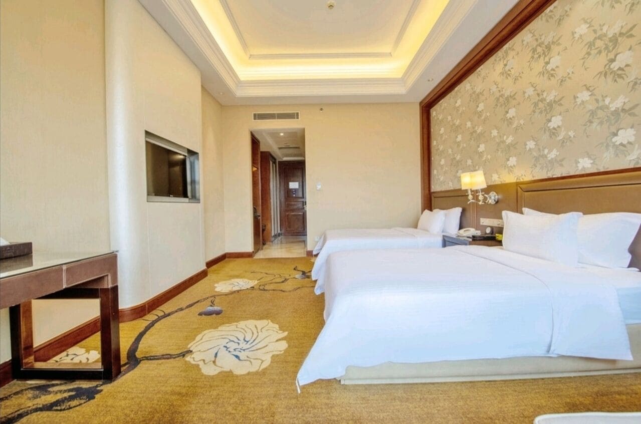 Habitación de hotel con dos camas, un escritorio, un televisor y una alfombra estampada con motivos florales.