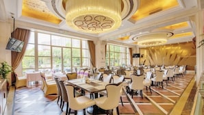A large banquet hall with multiple tables and chairs, large windows, and decorative chandeliers.