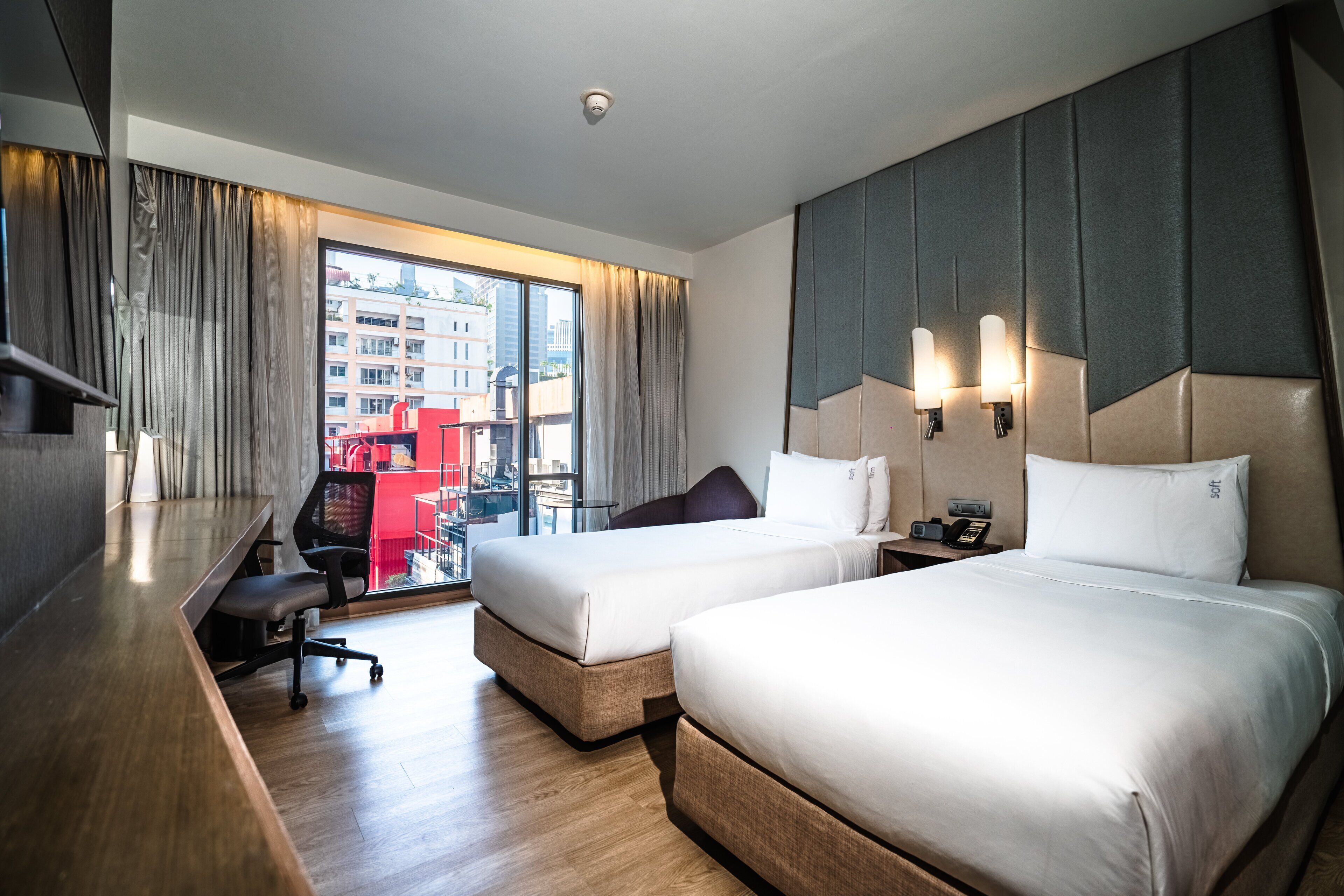 A hotel room with two beds, a desk, and a chair. There is a large window with curtains, a view of buildings outside, and a wall-mounted phone.
