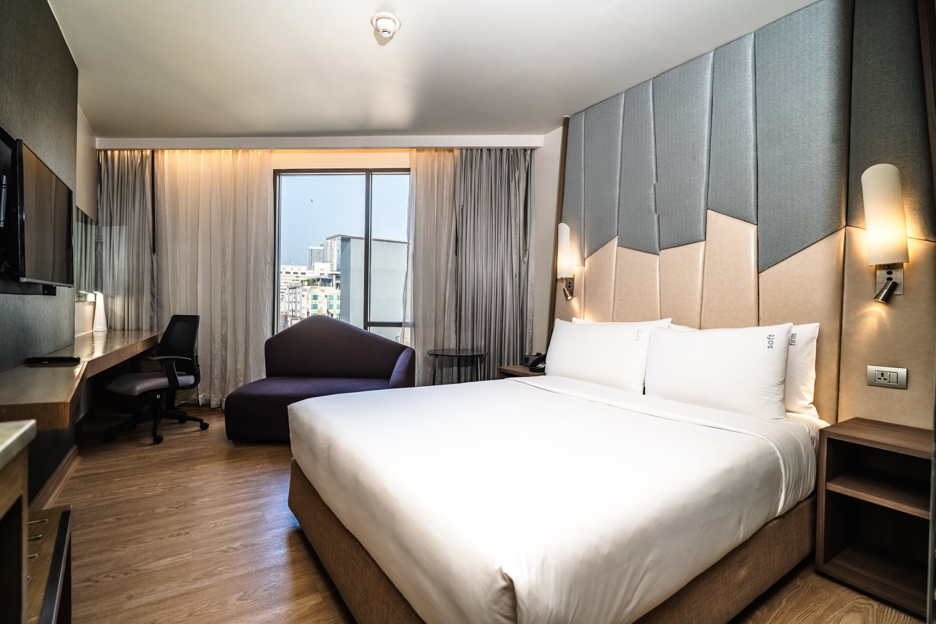 A hotel room with a large bed, a desk with a chair, a TV, and a view of the city through the window.
