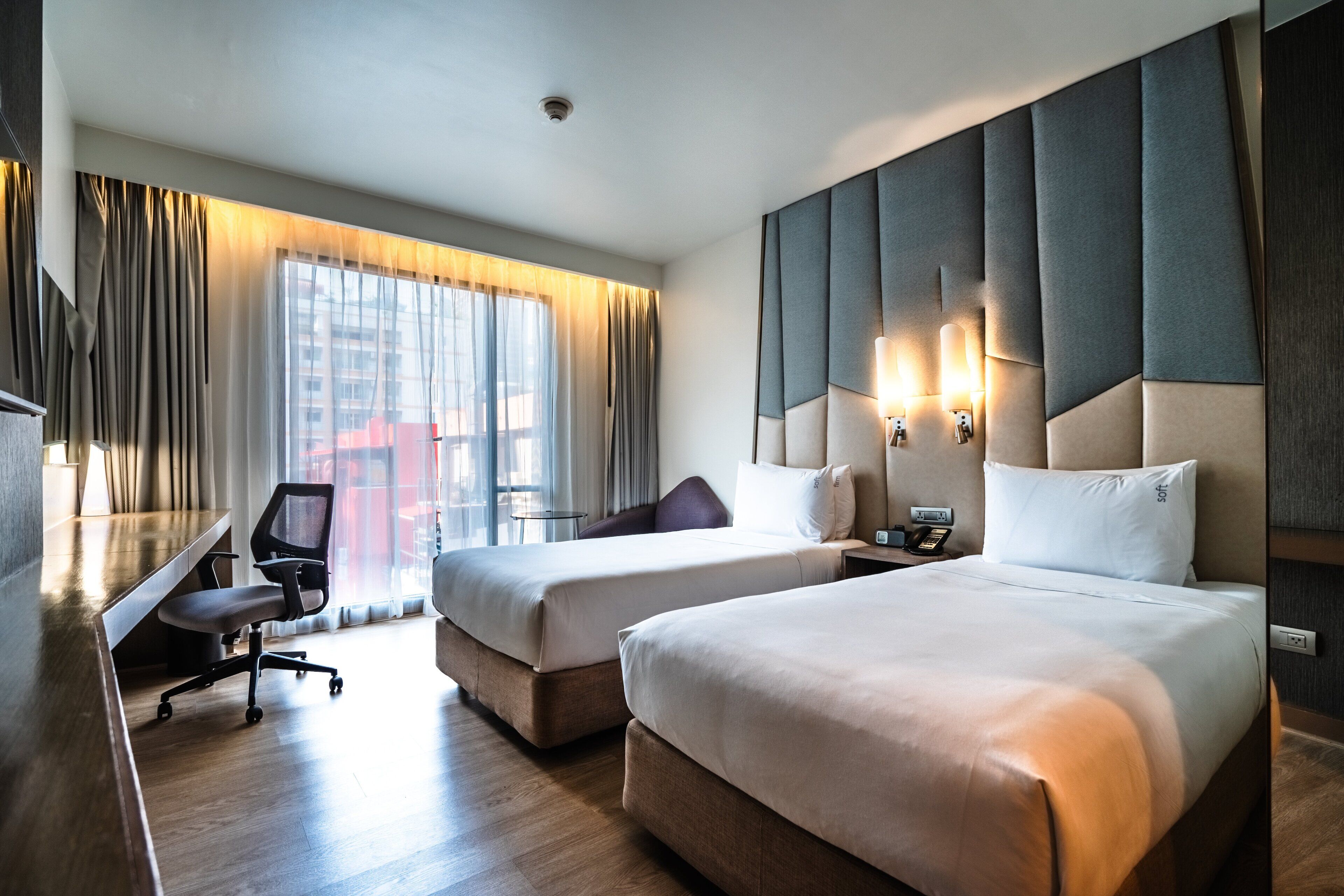 Hotelkamer met twee bedden, een bureau en een stoel. Groot raam met gordijnen, een wandlamp en een telefoon op het nachtkastje.