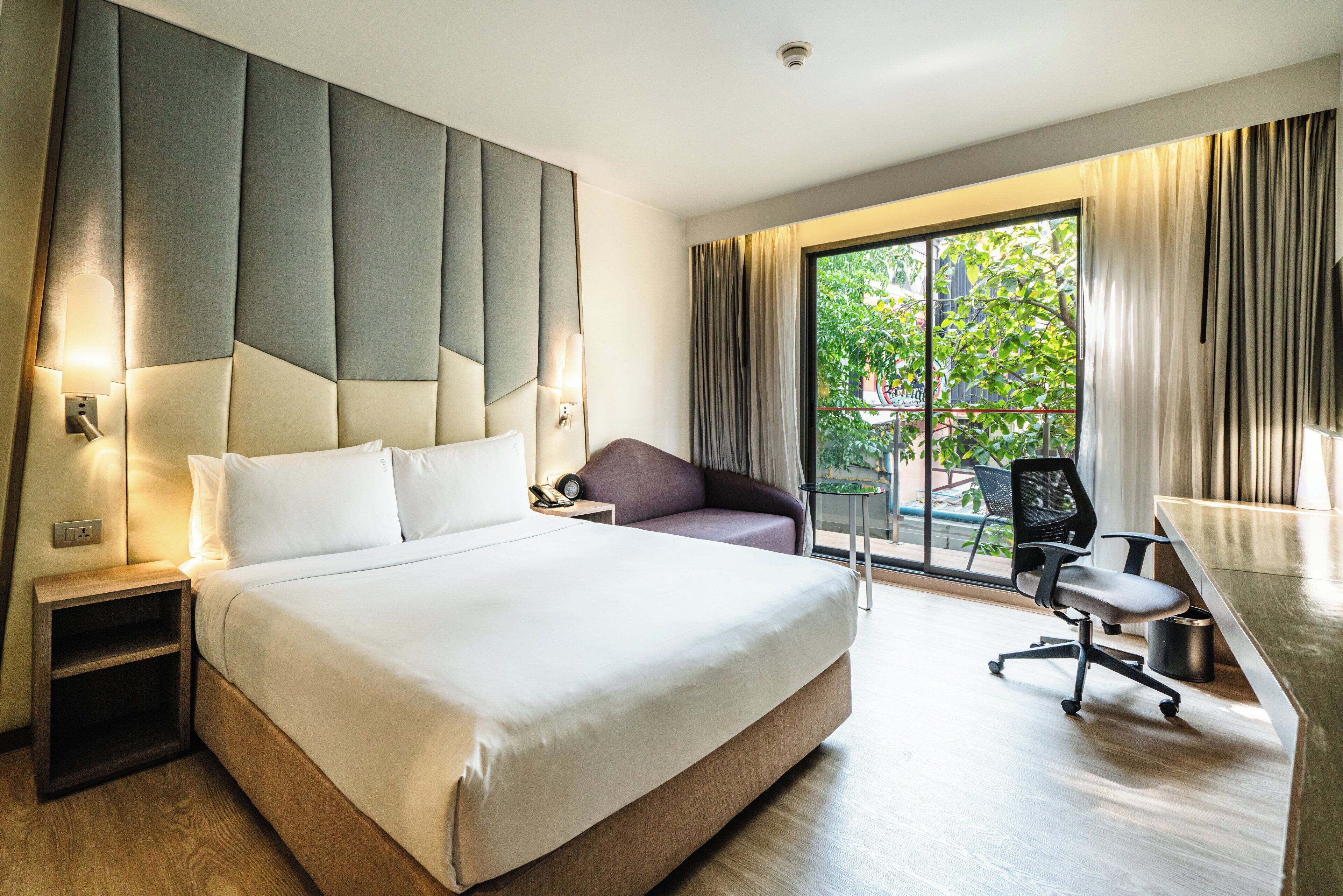 A hotel room with a large bed, a desk with a chair, a sofa, and a view of greenery outside.