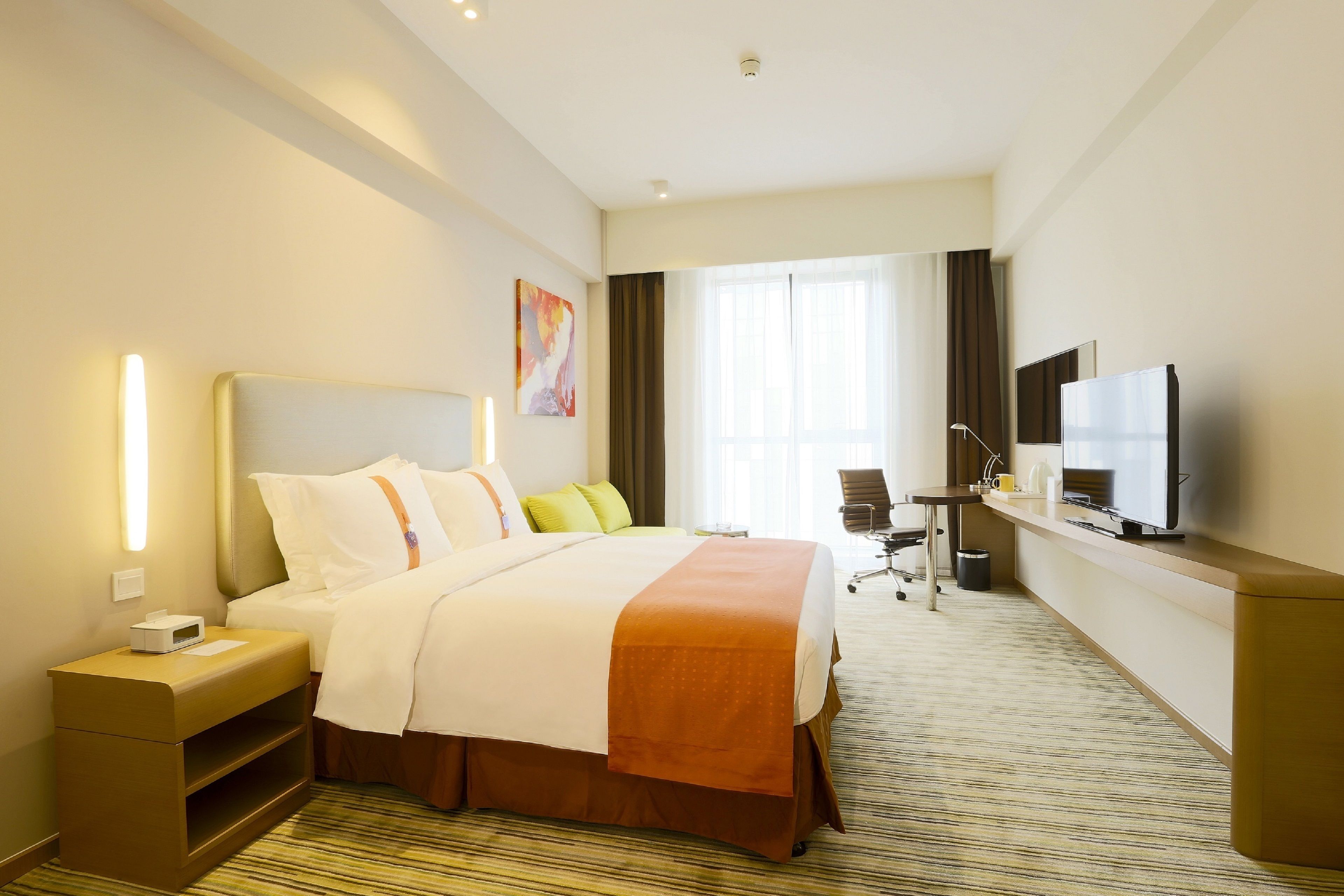 Habitación de hotel con una cama grande, un escritorio con televisor, una silla y un ventanal con cortinas.