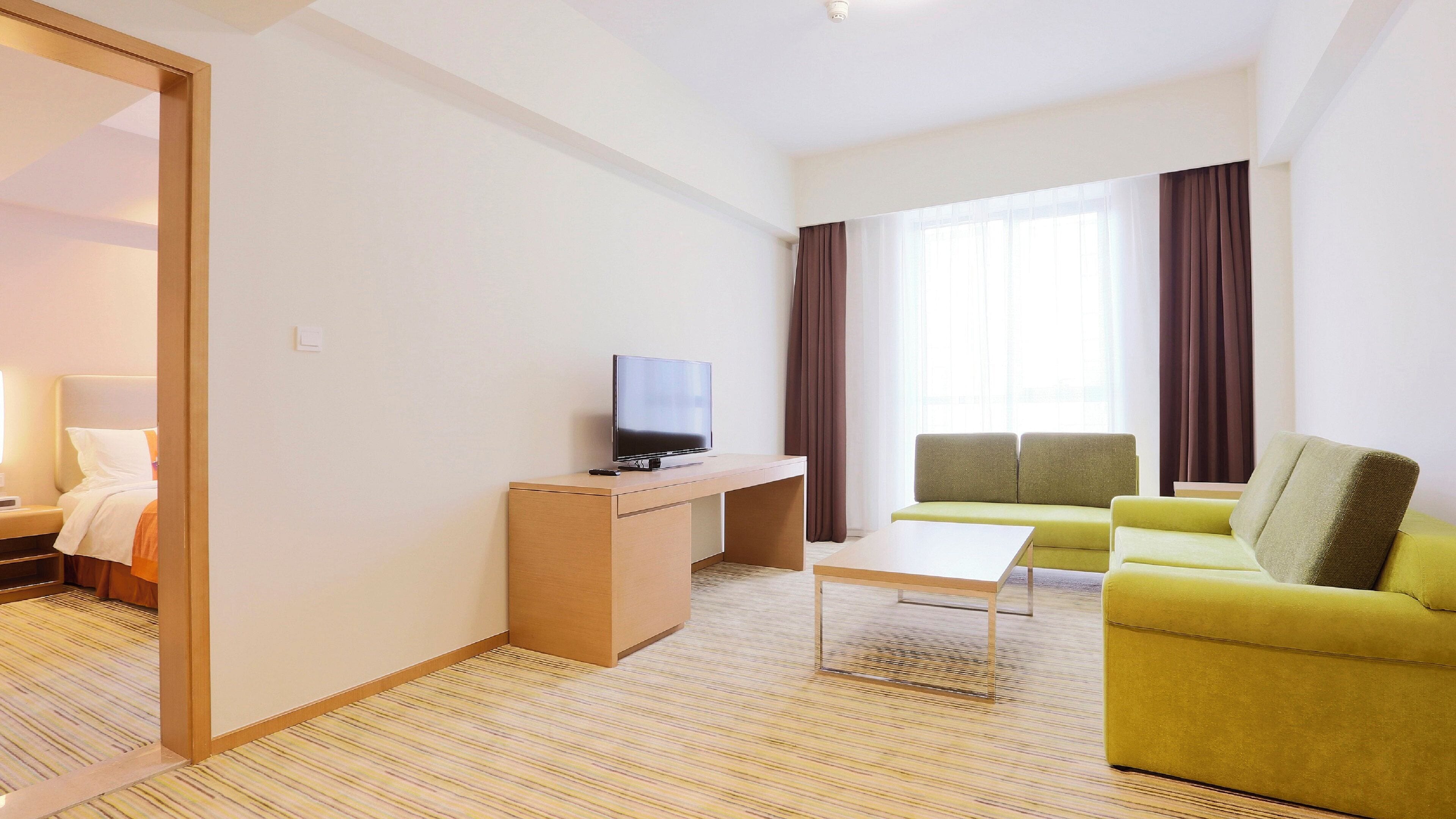 A hotel room with a bed, a TV on a desk, a sofa, and a window with curtains.