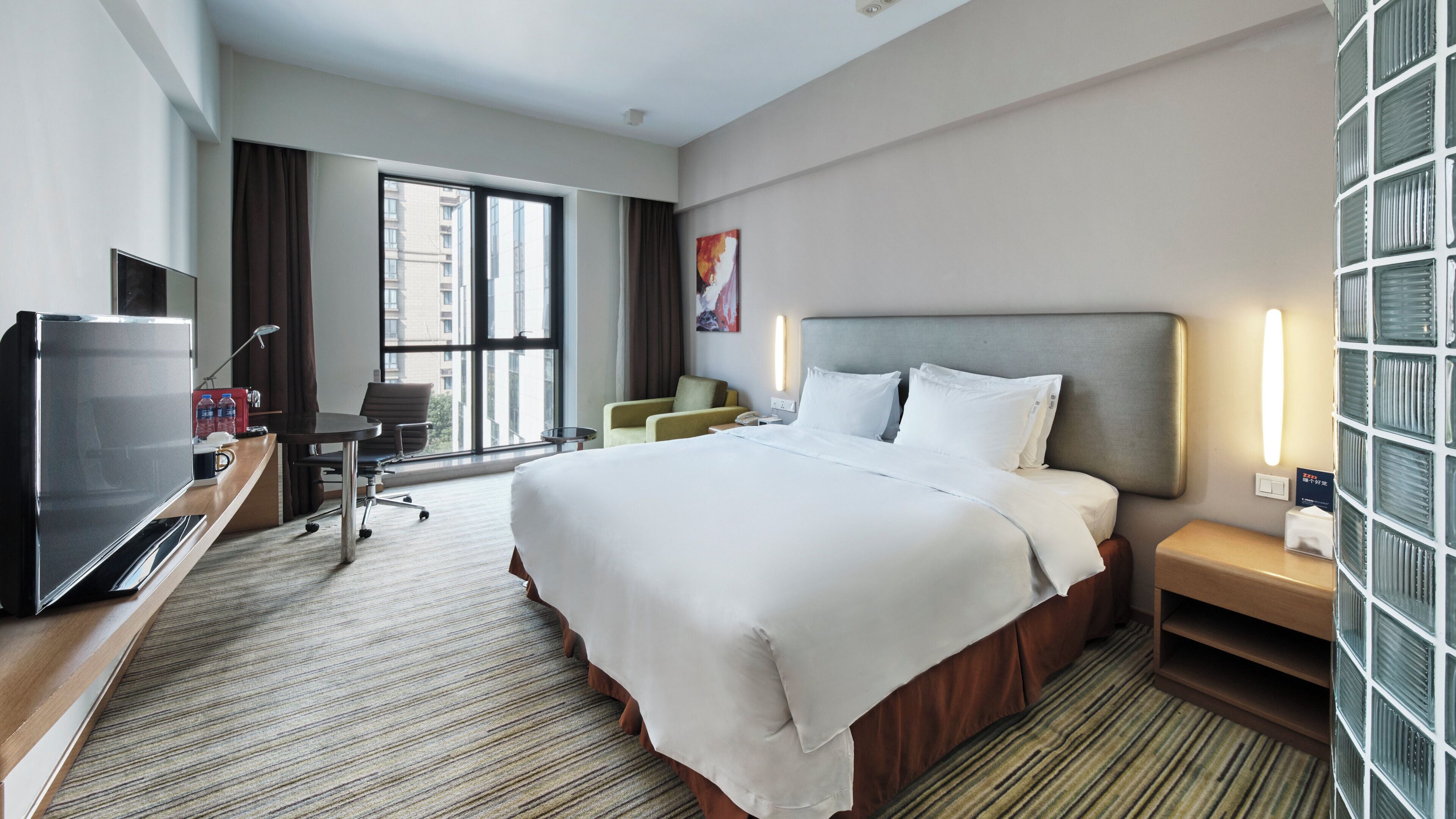 Een hotelkamer met een groot bed, een bureau met een televisie, een stoel, een bureaulamp en een raam met gordijnen.